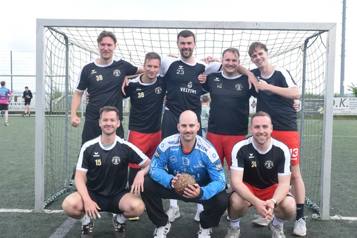 Eine Fußballmannschaft in schwarzen und roten Trikots posiert vor einem Fußballtor auf einem Kunstrasenplatz. Ein Torwart hält einen Ball fest.