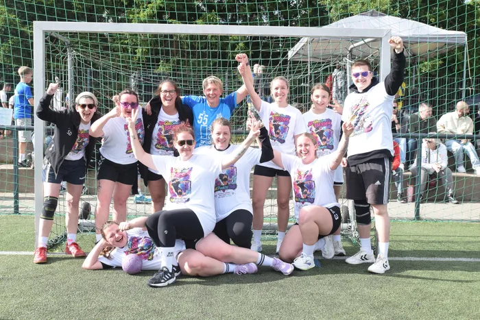 Eine Gruppe von jungen Frauen und Mädchen in Sportkleidung posiert jubelnd vor einem Handballtor. Sie tragen T-Shirts mit bunten Aufdrucken und zeigen ihre Freude. Im Hintergrund sind weitere Zuschauer und ein grüner Rasen zu sehen.
