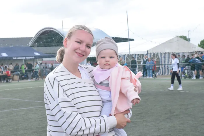 Eine junge Frau hält ein Baby im Arm und lächelt in die Kamera. Im Hintergrund ist ein Handballfeld und ein Stadion zu sehen.