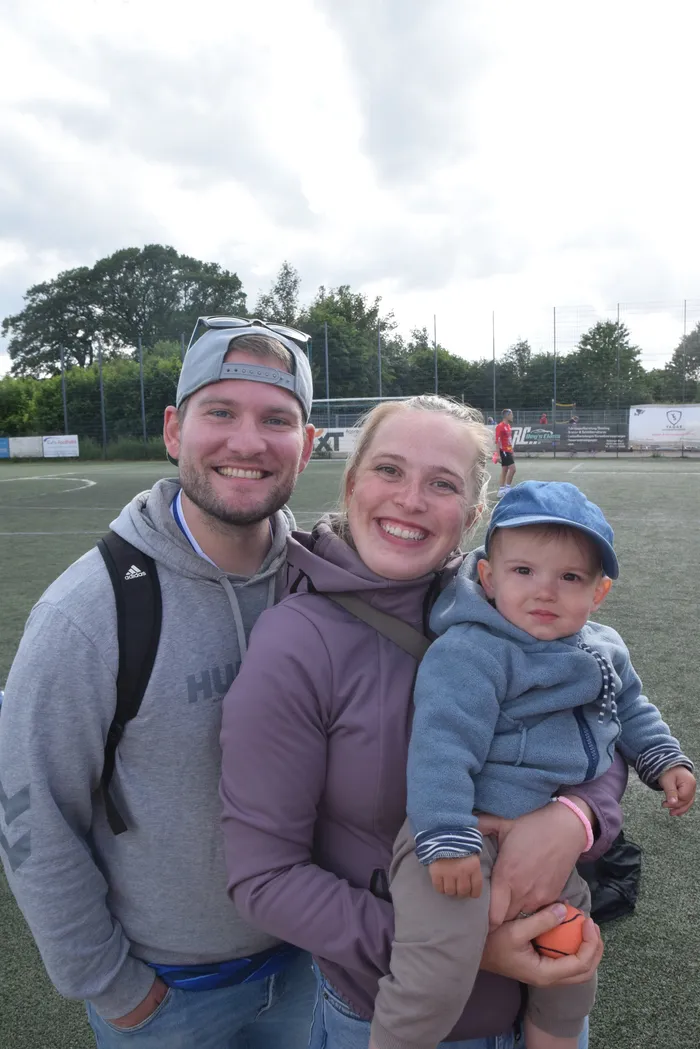 Ein lächelndes Paar steht mit einem kleinen Kind vor einer Sportanlage. Der Mann trägt eine Baseballkappe und einen Rucksack, die Frau hält das Kind auf dem Arm. Im Hintergrund ist ein Fußballfeld und eine Zuschauertribüne zu sehen.