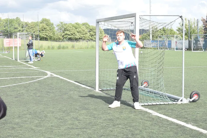 Ein junger Mann in Sportkleidung steht vor einem kleinen Handballtor auf einem grünen Rasenplatz. Im Hintergrund sind weitere Personen und Bäume zu sehen.