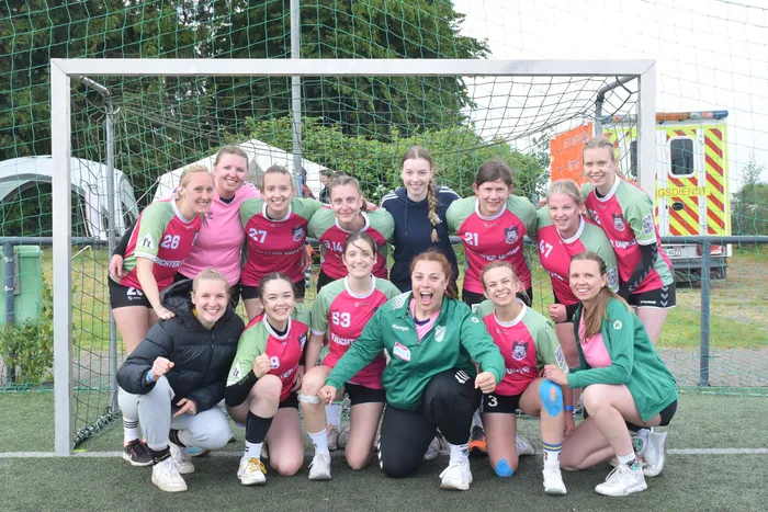 Eine Gruppe von jungen Frauen in Handballuniformen posiert vor einem Tor auf einem grünen Kunstrasenplatz. Einige tragen rote und pinke Trikots, andere grüne Trainingsjacken und schwarze Jacken.