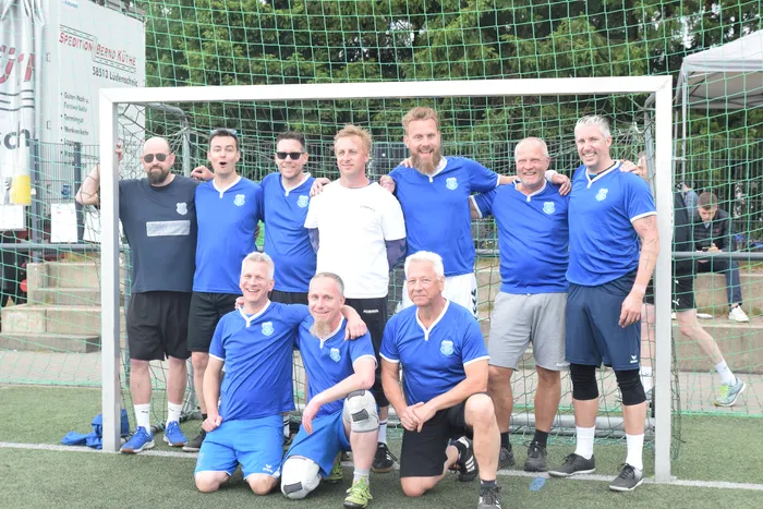 Eine Gruppe von Männern in blauen Sporttrikots und Shorts posiert vor einem Fußballtor auf einem Rasenplatz. Einige von ihnen lächeln in die Kamera, andere haben den Mund offen, als ob sie gerade jubeln. Im Hintergrund ist ein Gebäude mit einem Werbebanner zu sehen.
