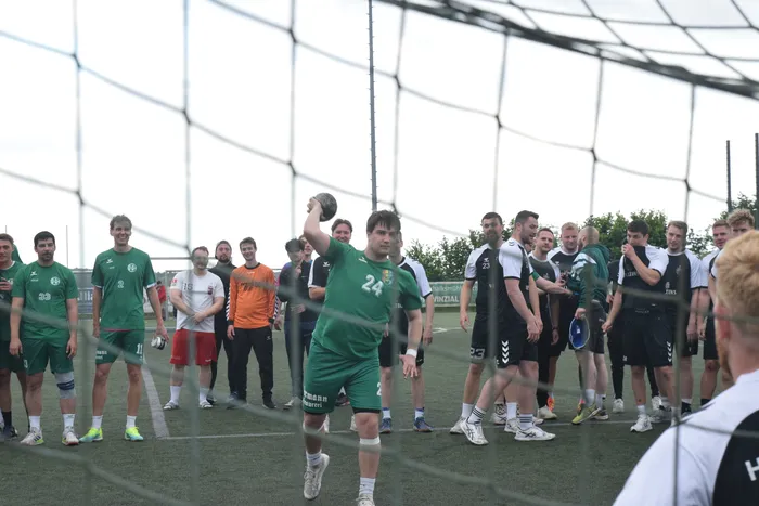Ein Handballspieler in grüner Ausrüstung wirft den Ball während eines Spiels auf dem Feld. Im Hintergrund stehen weitere Spieler und Zuschauer.