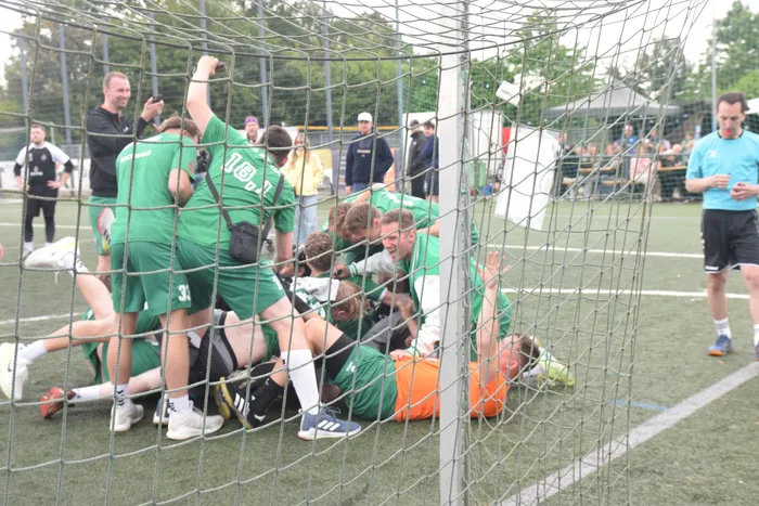 Eine Gruppe von Handballspielern in grünen und orangen Trikots feiert ausgelassen vor dem Tor. Im Hintergrund sind Zuschauer und weitere Spieler zu sehen.