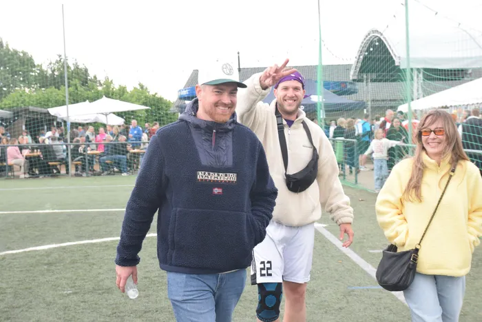 Drei Personen gehen auf einem Sportplatz, umgeben von Zuschauern und Zelten. Ein Mann mit Bart und Napapijri-Jacke lächelt, während ein anderer ihm zur Seite winkt. Eine Frau mit gelber Jacke geht neben ihnen.