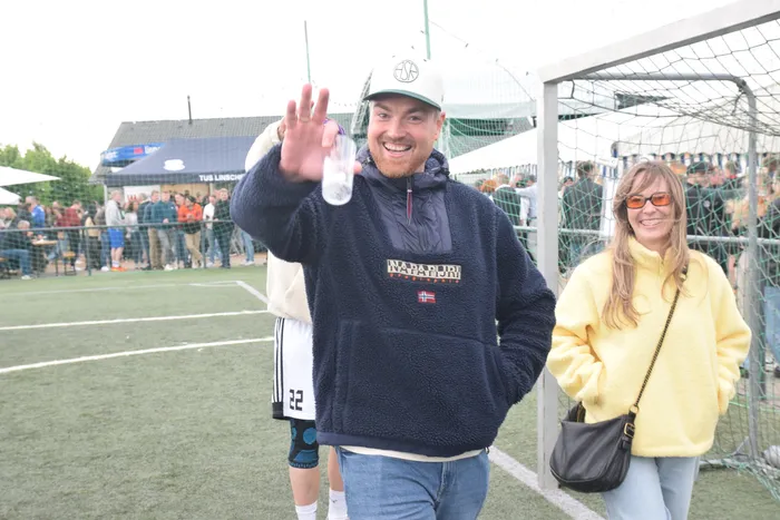 Ein lächelnder Mann mit hochgehobener Hand winkt in die Kamera. Neben ihm steht eine Frau und im Hintergrund sind Zuschauer auf einem Sportplatz zu sehen.