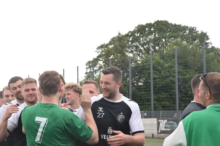 Feiernde Handballspieler umarmen sich und lächeln in der Nähe des Spielfelds, im Hintergrund Bäume und Zuschauer.