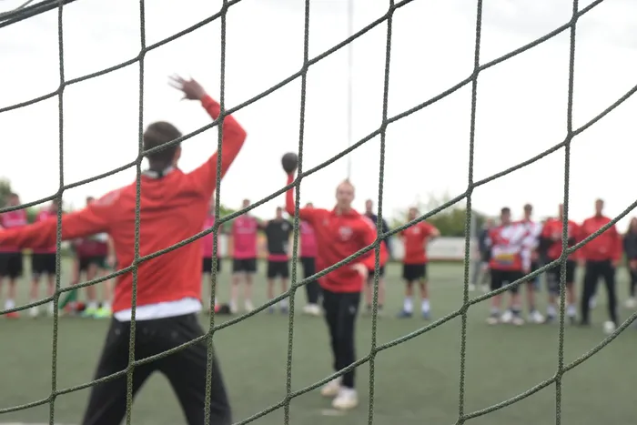 Handballspieler werfen und fangen Bälle auf einem grünen Feld, während sich im Hintergrund weitere Spieler und Zuschauer befinden. Das Bild ist durch ein Tornetz gerahmt.