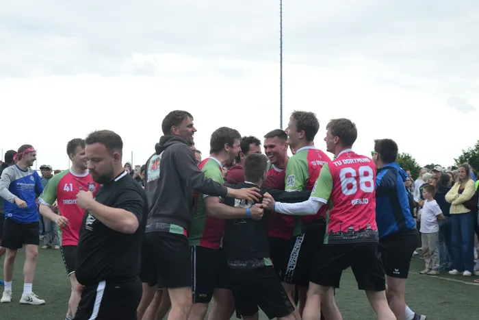 Eine Gruppe von jungen Männern in Sportkleidung umarmt und feiert auf einem grünen Feld. Im Hintergrund stehen Zuschauer und ein hoher Mast ist zu sehen.