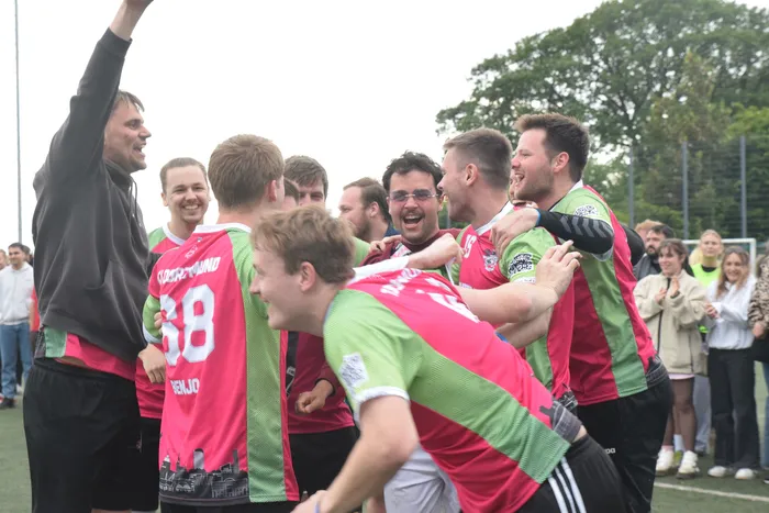 Eine Gruppe von Handballspielern in roten und grünen Trikots feiert ausgelassen, während ein Mann mit schwarzer Jacke den Arm in die Luft reckt. Im Hintergrund stehen Zuschauer an einem Sportplatz.