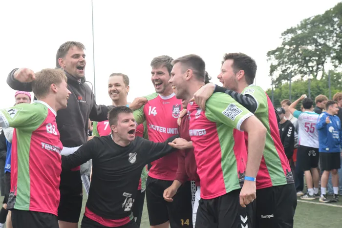 Feiernde Handballspieler umarmen sich und jubeln auf dem Spielfeld. Im Hintergrund sind weitere Spieler und Zuschauer zu sehen.