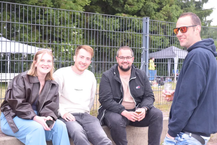 Vier lächelnde Menschen sitzen oder stehen vor einem Maschendrahtzaun und halten Getränke in der Hand. Im Hintergrund sind weitere Personen und Zelte zu sehen.