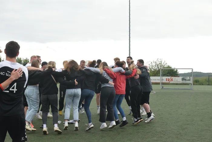 Eine Gruppe von jungen Erwachsenen steht eng umschlungen auf einem grünen Sportplatz, vermutlich nach einem Handballspiel. Im Hintergrund sind Tornetze und ein Mast zu erkennen.