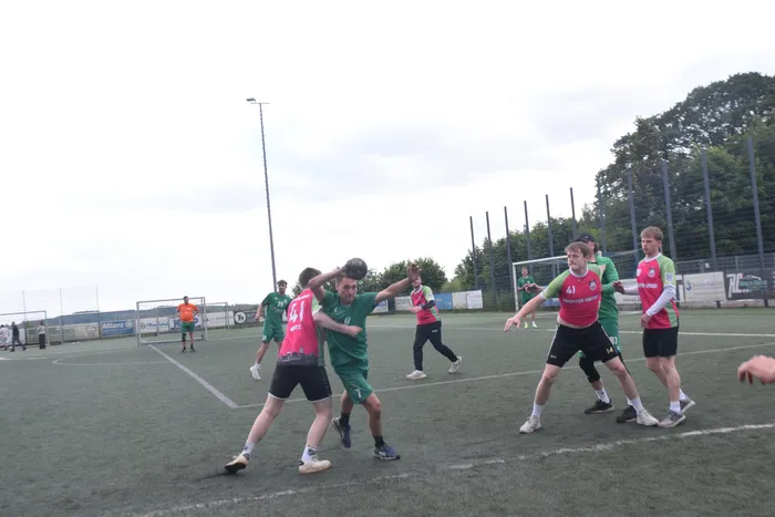 Fußballspieler in grünen und roten Trikots kämpfen um den Ball auf einem Kunstrasenplatz. Im Hintergrund sind weitere Spieler und Zuschauer zu sehen.
