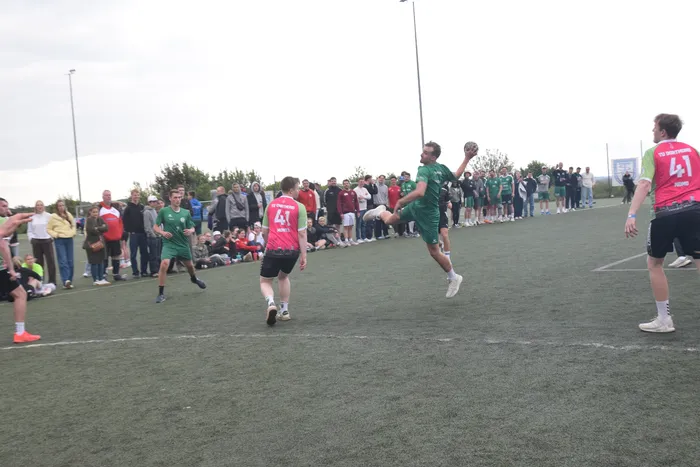 Ein Handballspieler in Grün springt zum Wurf, während ein weiterer Spieler in Pink bereit steht. Im Hintergrund stehen Zuschauer auf einem Sportplatz.