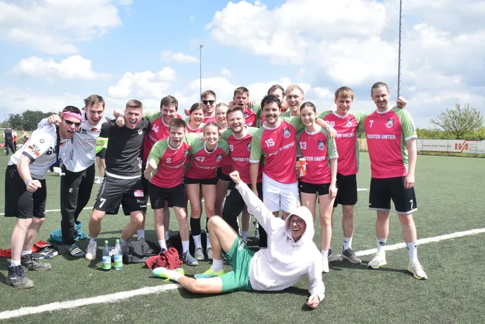 Eine Gruppe von jungen Erwachsenen in Sportkleidung posiert auf einem grünen Kunstrasenfeld. Viele tragen rote Trikots mit der Aufschrift „Enniger United“, während ein Mann in einer weißen Trainingsjacke und Hose im Vordergrund liegt und jubelt.