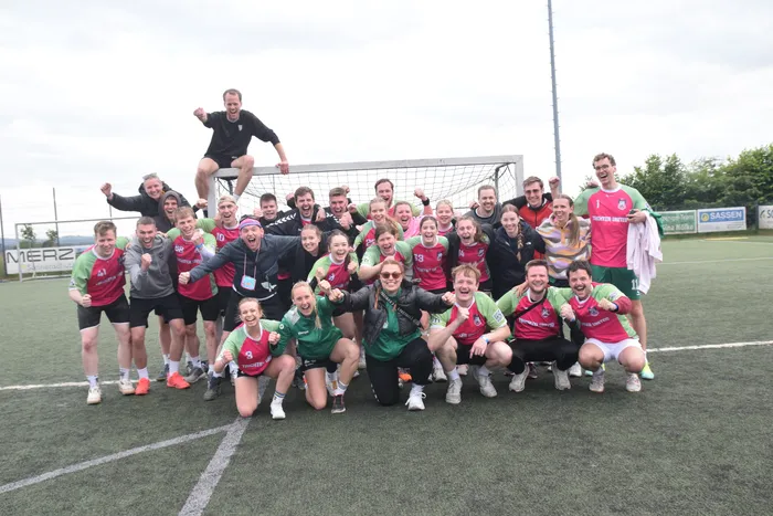 Eine Gruppe von etwa 20 Personen, hauptsächlich junge Erwachsene, posiert lächelnd auf einem Kunstrasenplatz vor einem Fußballtor. Einige tragen rote und grüne Trikots, andere dunkle Kleidung. Ein Mann steht auf dem Tornetz und jubelt.