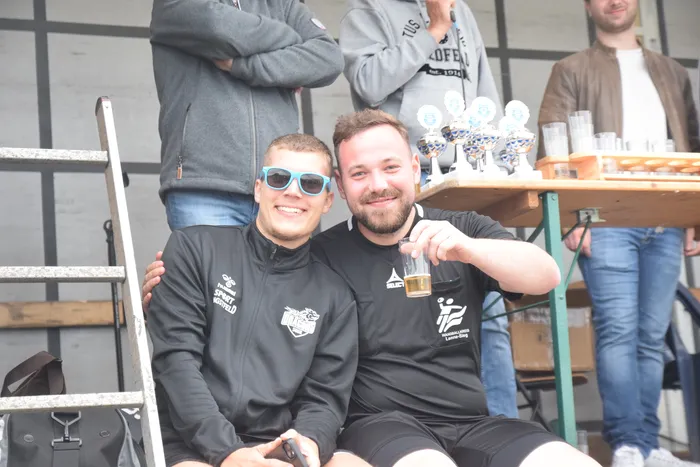Zwei lächelnde Männer in dunklen Sportjacken sitzen nebeneinander, einer hält ein Bierglas. Im Hintergrund stehen Trophäen auf einem Tisch und weitere Personen.