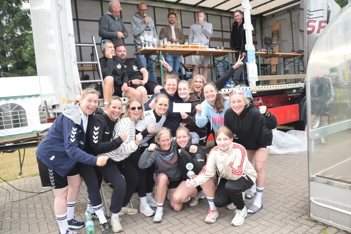 Eine Gruppe von etwa 20 Personen, darunter viele junge Frauen in Sportkleidung, posiert vor einem Lastwagen, der als Bühne dient. Einige halten Trophäen oder Urkunden in die Höhe und lächeln in die Kamera. Im Hintergrund ist ein Zelt und Bäume zu sehen.