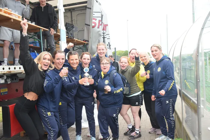 Eine Gruppe von Handballspielerinnen in blauen Trainingsanzügen posiert mit einem Pokal und Bierflaschen vor einem weißen Transporter und einer Gewächshauskonstruktion. Sie lächeln und zeigen ihre Freude über den Gewinn.