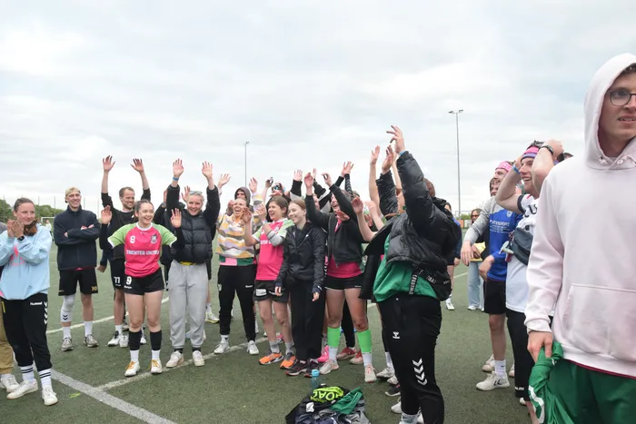 Eine Gruppe von Frauen und Männern steht auf einem Rasenplatz und hebt die Hände in die Luft. Viele tragen Sportkleidung, einige Trikots mit der Aufschrift 