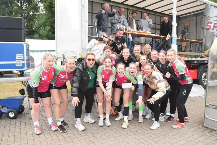 Eine Gruppe von jungen Frauen in Handballuniformen posiert eng zusammen und hält einen Pokal hoch. Im Hintergrund stehen und jubeln weitere Personen, darunter auch Männer, vor einem weißen Transporter.