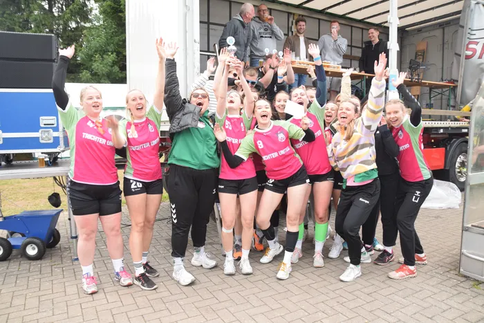 Eine Gruppe von Handballspielerinnen in rosa und grünen Trikots jubelt mit erhobenen Armen und feiert gemeinsam. Im Hintergrund stehen und jubeln weitere Personen auf einem Anhänger.