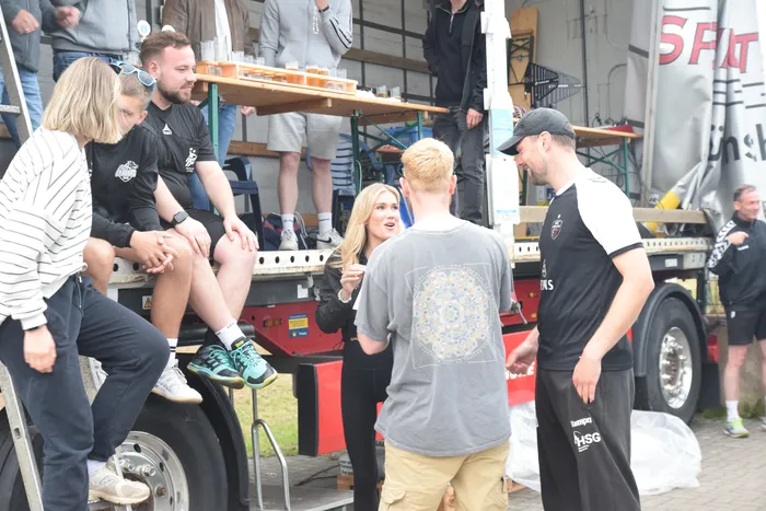 Menschen stehen und sitzen auf und um einen Anhänger, der als provisorische Theke dient. Im Hintergrund sind weitere Personen und ein Fahrzeug mit Werbeaufdrucken zu sehen.