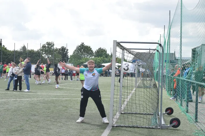 Ein Jugendlicher steht mit ausgebreiteten Armen vor einem Handballtor auf einem grünen Rasenplatz. Im Hintergrund sind weitere Personen und ein grünes Netztor zu sehen.