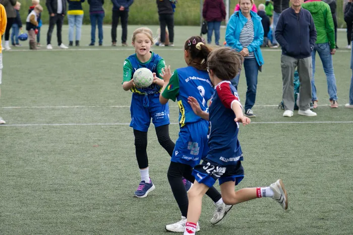 Zwei junge Fußballspielerinnen in blauen Trikots und schwarzen Shorts stehen auf einem grünen Rasenfeld. Die Spielerin links hält einen Ball, während die rechts in Laufhaltung ist. Im Hintergrund stehen Zuschauer.