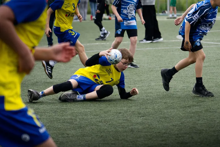 Ein Jugendlicher in gelbem Trikot und blauen Shorts rutscht auf dem grünen Kunstrasen und hält einen Ball fest, während ein Gegenspieler in blauem Trikot und Shorts auf ihn zuläuft. Im Hintergrund stehen weitere Spieler und Zuschauer.