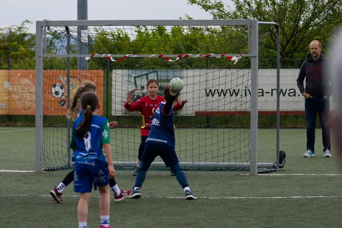 Zwei junge Fußballspielerinnen stehen vor einem Tor und bereiten sich auf einen Schuss vor, während ein Jugendlicher im Hintergrund den Ball hält. Ein Mann beobachtet das Spiel am Spielfeldrand.