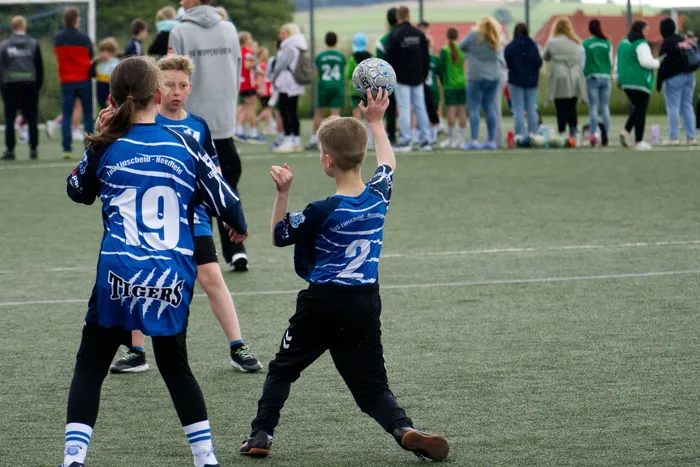 Zwei junge Fußballspieler im blauen Trikot mit der Aufschrift „Tigers“ und den Nummern 19 und 2 stehen auf einem grünen Rasenfeld. Der Spieler mit der Nummer 2 wirft einen Ball in die Luft, während die Spielerin mit der Nummer 19 zusieht. Im Hintergrund sind weitere Spieler und Zuschauer zu erkennen.