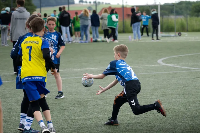 Ein Jugendlicher in blauem Trikot mit der Nummer 2 versucht, den Ball einem Gegenspieler in gelbem Trikot mit der Nummer 7 abzunehmen. Im Hintergrund stehen weitere Kinder und Jugendliche auf einem Sportplatz.