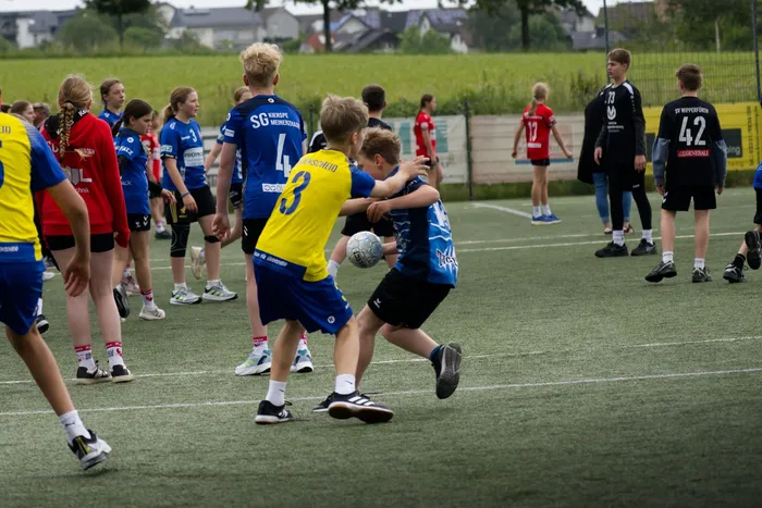 Zwei Fußballspieler kämpfen um den Ball auf einem Kunstrasenplatz, umgeben von weiteren Spielern und Zuschauern. Im Hintergrund sind Bäume und Gebäude zu sehen.