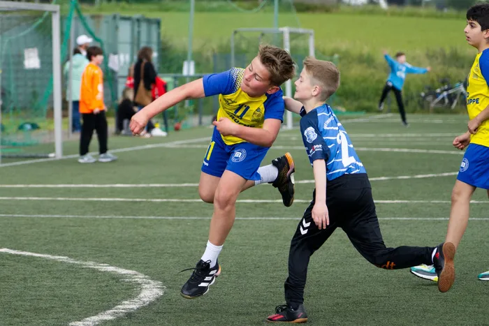 Zwei Fußballspieler, ein Junge in Blau und Schwarz und ein anderer in Gelb und Blau, kämpfen um den Ball auf einem Kunstrasenplatz. Im Hintergrund stehen weitere Spieler und Zuschauer.