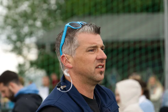 Ein Mann mit blauen Sonnenbrillen und Headset spricht in die Ferne, im Hintergrund verschwommene Menschen und Grünflächen.