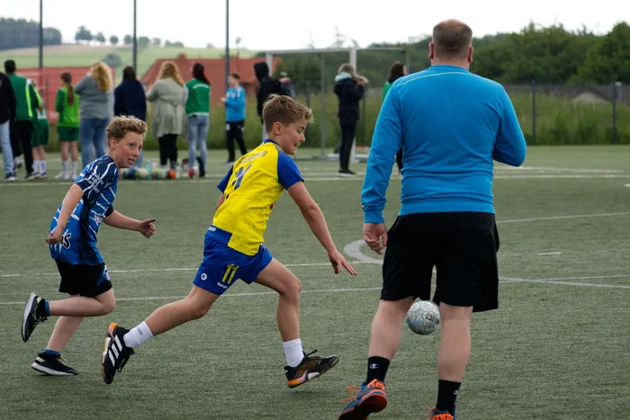Zwei junge Fußballspieler in Vereinskleidung dribbeln den Ball auf einem Kunstrasenplatz, während ein Erwachsener im Hintergrund zuschaut. Im Hintergrund stehen weitere Personen und Gebäude.