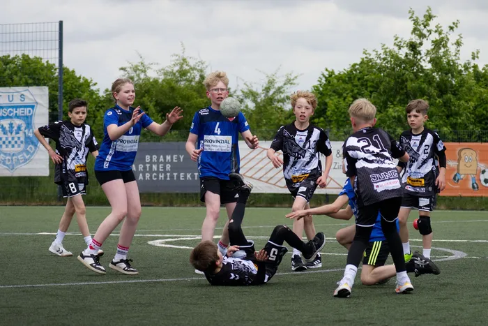 Jugendliche Fußballspieler in blauen und schwarz-weißen Trikots kämpfen um den Ball auf einem Kunstrasenplatz. Ein Spieler liegt im Vordergrund, während andere um ihn herum stehen und zum Ball greifen. Im Hintergrund ist eine grüne Hecke und ein Zaun mit Werbeplakaten zu sehen.