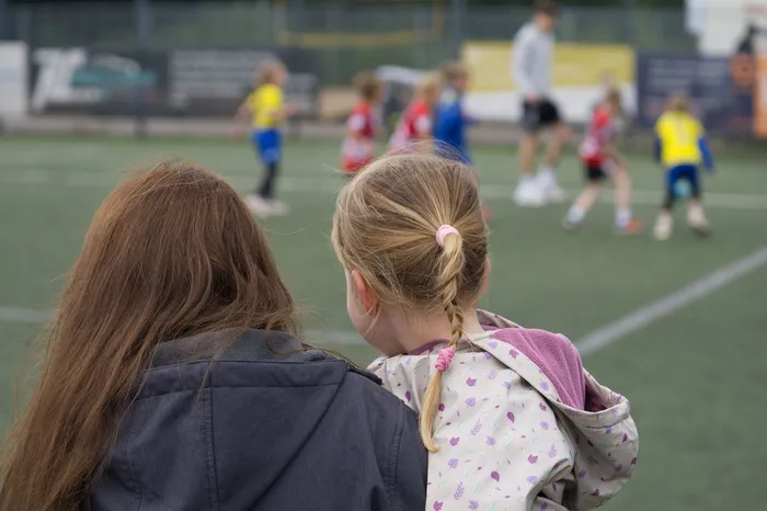 Zwei Mädchen mit langen Haaren sitzen einander zugewandt und schauen auf ein Fußballspiel im Hintergrund. Kinder in roten und gelben Trikots spielen auf einem grünen Rasenfeld.