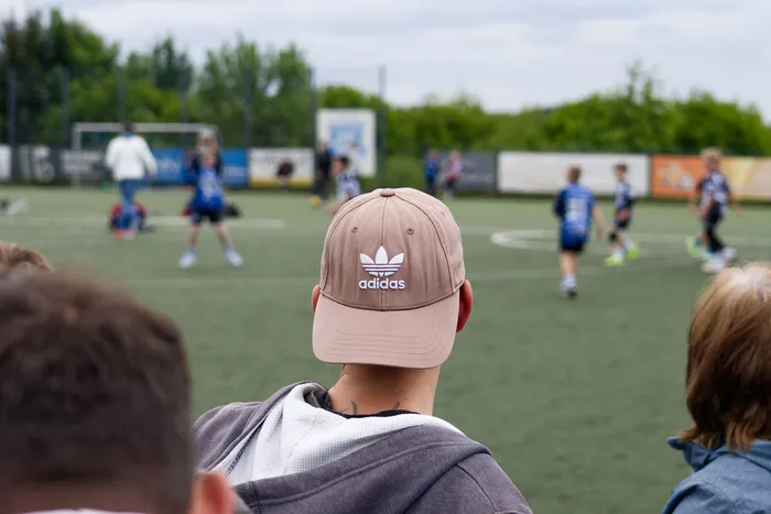 Zuschauer sitzen auf einer Bank und beobachten ein Fußballspiel mit Kindern und Jugendlichen auf einem Kunstrasenplatz. Im Vordergrund ist der Hinterkopf einer Person mit einer beigen Adidas-Kappe zu sehen.