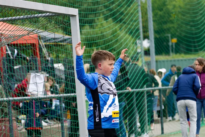 Ein Jugendlicher steht vor einem Fußballtor und hebt die Arme. Er trägt einen blauen Trainingsanzug mit dem Aufdruck eines Wolfskopfes und Sponsorenlogos. Im Hintergrund sind weitere Personen und ein Zaun zu sehen.