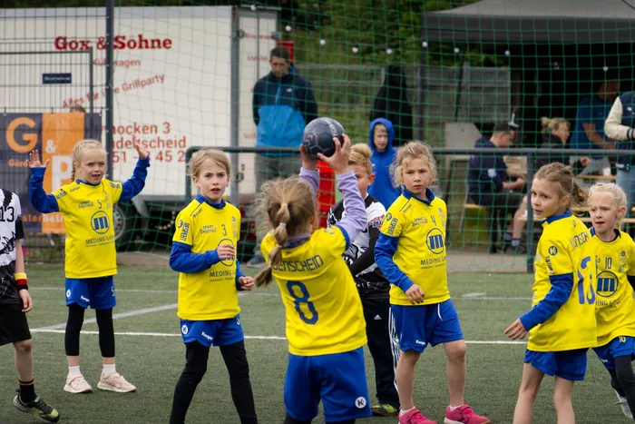 Eine Gruppe von Kindern in Fußballtrikots steht auf einem Kunstrasenplatz. Einige jubeln mit erhobenen Armen, während andere aufmerksam zusehen. Im Hintergrund sind weitere Kinder und ein Zaun zu erkennen.