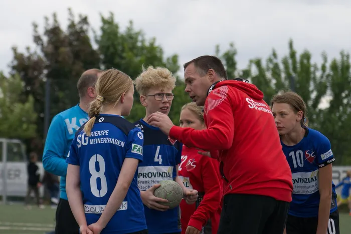 Ein Trainer in roter Trainingsjacke gibt mehreren jungen Fußballspielerinnen Anweisungen. Die Spielerinnen tragen blaue und gelbe Trikots mit Vereinsaufschriften. Im Hintergrund sind Bäume und ein Fußballtor zu sehen.
