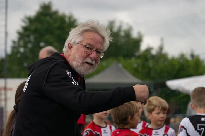 Ein älterer Mann mit Bart und Brille gestikuliert energisch vor einer Gruppe Jugendlicher in roten Trikots. Im Hintergrund sind Bäume und ein weißes Zelt zu sehen.