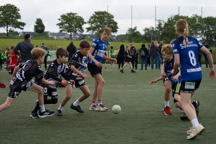 Eine Jugendfußballmannschaft in schwarzen und weißen Trikots kämpft um den Ball gegen eine Mannschaft in blauen Trikots auf einem Kunstrasenplatz. Im Hintergrund stehen Zuschauer und weitere Spieler.