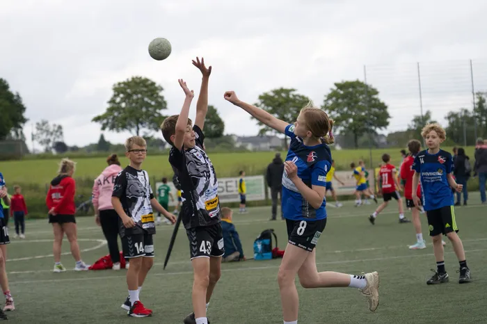 Zwei Jugendliche springen auf einem Sportplatz in der Luft, um einen Ball zu erreichen. Im Hintergrund sind weitere spielende Kinder und Zuschauer zu sehen.