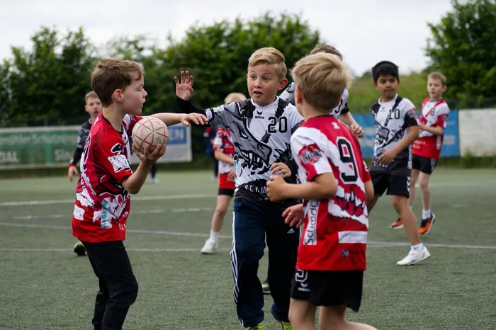 Junge Fußballspieler in Rot und Schwarz stehen auf einem grünen Rasenplatz und spielen miteinander. Ein Spieler wirft den Ball einem anderen zu. Im Hintergrund weitere Kinder beim Spiel.