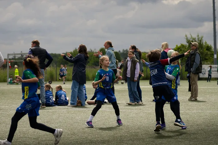 Junge Fußballspielerinnen und Fußballspieler in blauen und schwarzen Trikots spielen auf einem grünen Kunstrasenplatz, umgeben von Zuschauern. Einige Zuschauer stehen und applaudieren.